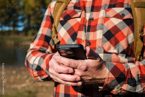 Man in plaid shirt using smartphone by lake during early autumn, holding mobile device outdoors against peaceful natural landscape, digital communication concept