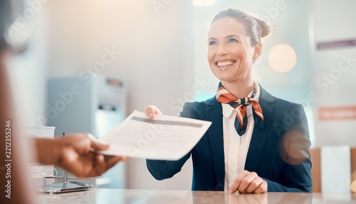 Smiling female customer service agent in uniform assists a passenger with travel documents at the reception counter