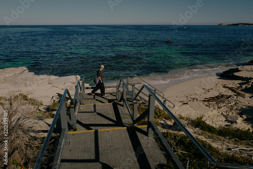 Bald man descending steps toward a sandy beach with clear blue w