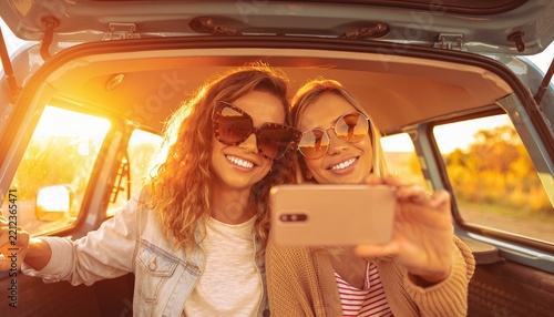 Two happy young women taking a selfie with a smartphone while sitting in the trunk of a car during a sunny road trip adventure at sunset