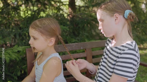 Young girl braids sister hair outdoors while seated on wooden garden bench, showing sibling conflict annoyance as younger child sits with bored impatient expression in sunny park setting.