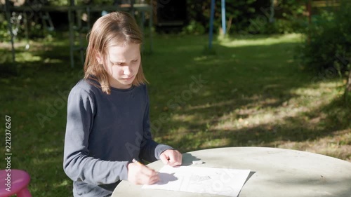 Young girl draws with pencil on paper at round stone table in sunny garden. Focused female child sketches outdoors during daytime, displaying thoughtful emotion while sitting on stool.
