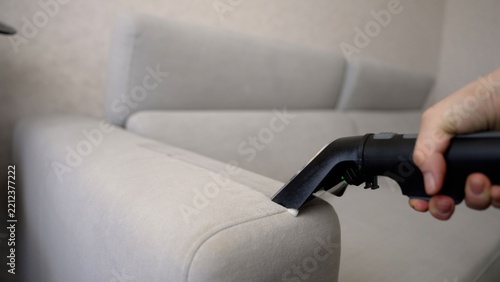 Black vacuum cleaner nozzle in hand cleaning light grey fabric sofa armrest, professional upholstery maintenance