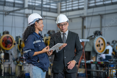 A female factory worker and male executive wearing safety helmets discuss production operations using a tablet inside an automotive parts manufacturing plant.