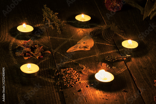 Witchy altar with drawn pentagram circle decorated burning candles, a bird skull, dried herbs, autumn flowers, crystals and an old key on wooden surface in the dark