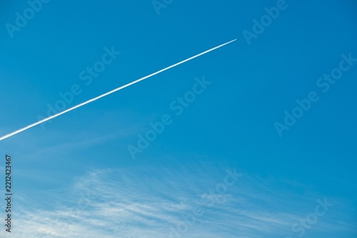 Inversion contrail of an airplane cutting through a clear blue sky with light wispy clouds.