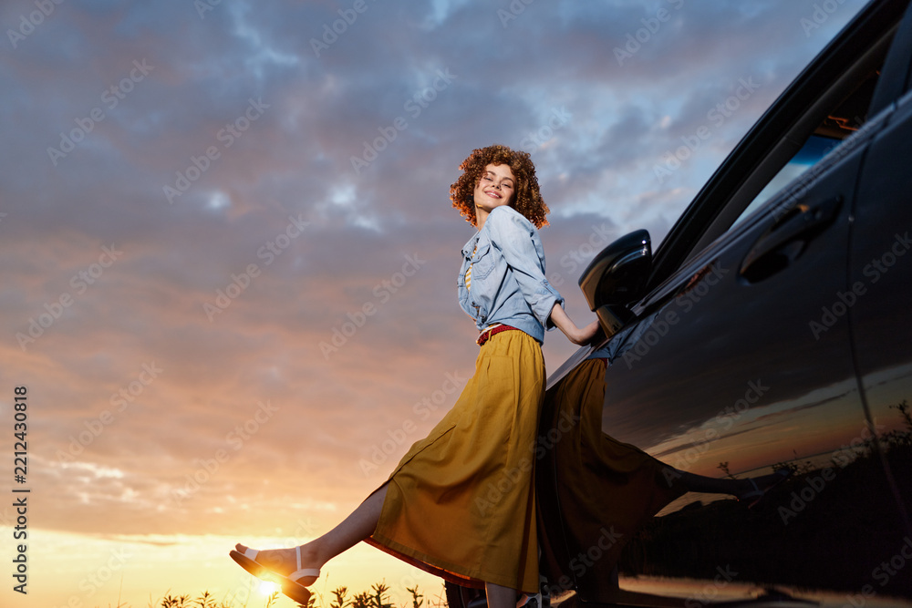 Obraz premium Woman lifestyle scene with a joyful adult woman by a car at sunset, wearing a yellow skirt and denim shirt, leaping in a carefree pose against a warm sky.