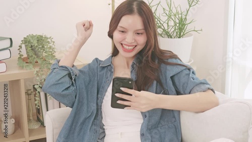 A happy young Asian woman sitting on a sofa and using a smartphone. She smiles and celebrates a success.