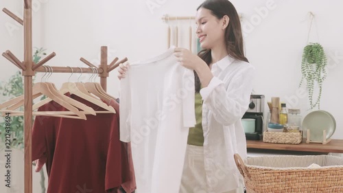 A smiling young Asian woman folding a t-shirt and organizing clothes into a laundry basket at home.