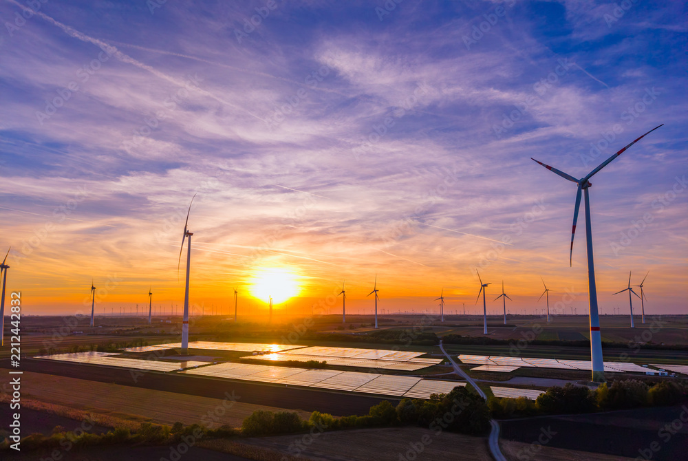 Obraz premium Wide aerial panorama of wind turbine farm and solar collectors at sunset. Colorful evening sky illuminates agricultural land filled with green power installations.