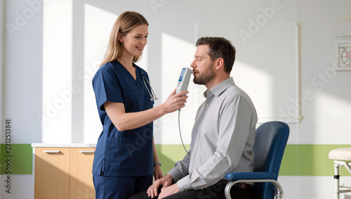 A nurse uses a contactless thermometer to check a male patient's temperature before his appointment during pre-appointment screening and clinic intake at a medical reception