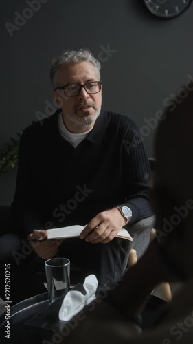 Vertical shot of middle aged Caucasian psychologist writing notes while listening to client during therapy session. Suitable for mental healthcare and medical projects