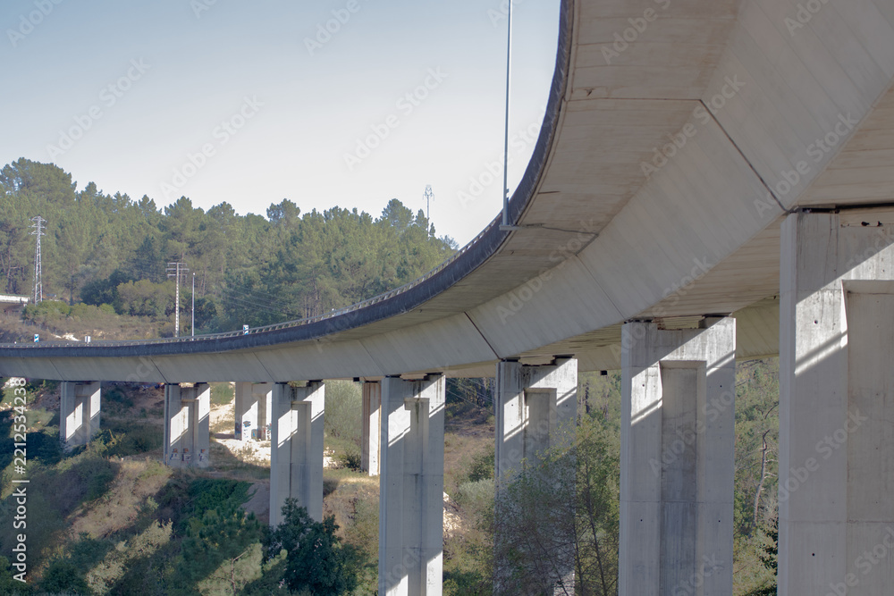 Fototapeta premium Curved concrete highway viaduct with tall support pillars over a wooded valley, low angle view under the bridge with clear blue sky