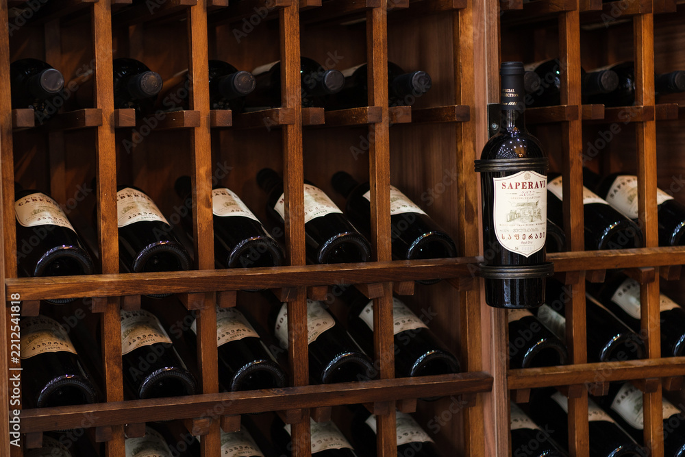 Naklejka premium Patardzeuli, Georgia - September 03, 2026: Wooden rack holds rows of dark wine bottles, with a labeled saperavi bottle displayed prominently among cellar storage