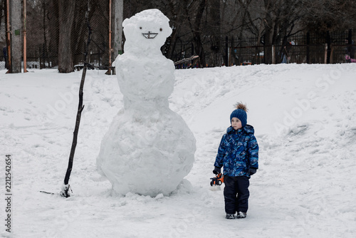 Little boy in blue winter jacket and hat stands next to a huge giant snowman outdoors in snow covered park on a winter day