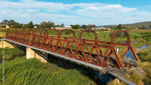 Aerial view of an abandoned rusted railway bridge spanning a river with overgrown vegetation and surrounding rural landscape in Oudtshoorn, Western Cape, South Africa.