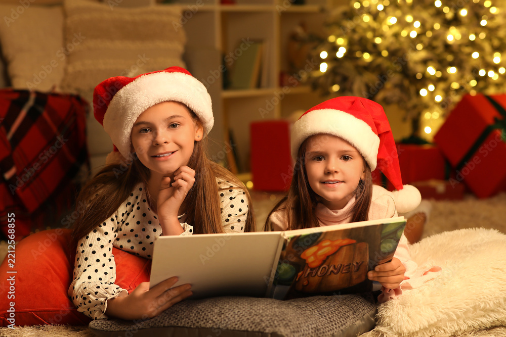 Obraz premium Cute happy little girls in Santa hats with book reading Christmas story at night