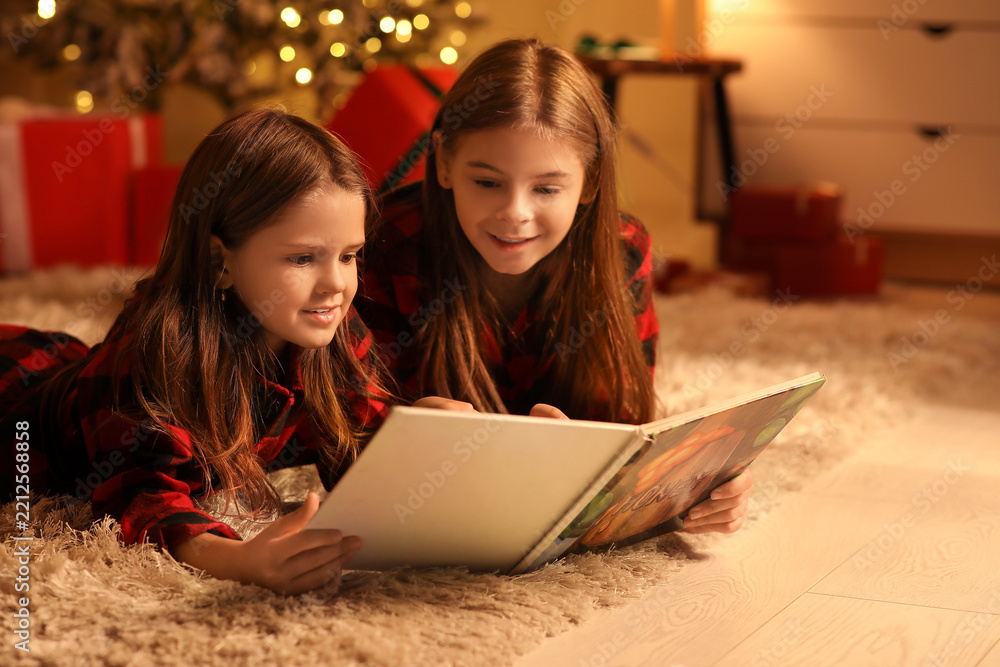 Obraz premium Cute happy little girls in checkered pajamas with book reading Christmas story at night