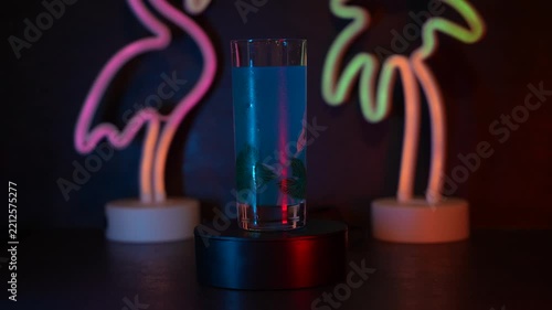Blue glass rotating 360 degrees on a dark pedestal with colorful neon flamingo and palm tree lights in the background. Vibrant tropical nightlife atmosphere.