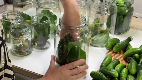 a person puts green cucumbers into a glass jar, multiple glass jars with leaves stand on a white table, a wooden board holds more cucumbers