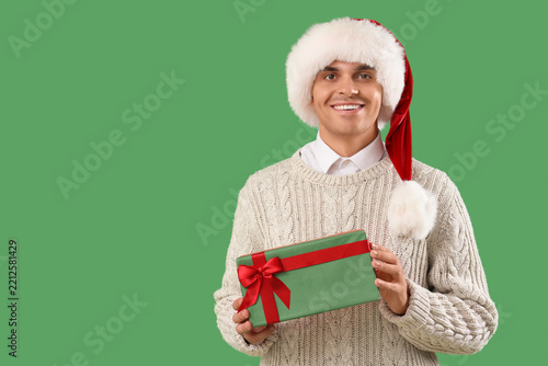 Happy young man in Santa hat with gift box on green background