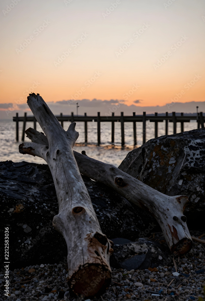 Fototapeta premium Serenity and mindfulness, driftwood on a rocky beach at sunset with a wooden pier in the background for travel and coastal nature relaxation concepts