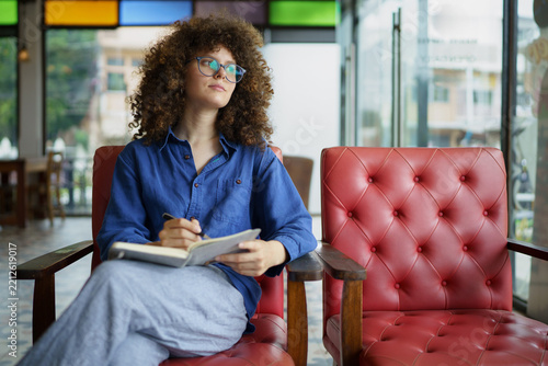 A focused young woman with curly hair and glasses writes in a notebook while sitting in a vibrant cafe, ideal for creativity and lifestyle concepts