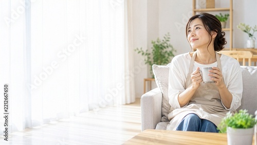 明るいリビングで飲み物を手にくつろぐ30代の日本人女性、家事の合間にひと息ついてリラックスする暮らしのイメージ