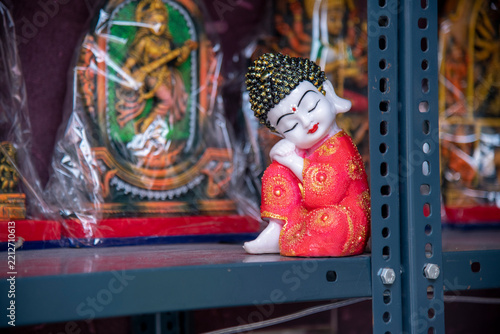 Close-up shot of a small sleeping monk Buddha statue wearing a bright red floral robe sitting peacefully on a metal shop shelf, surrounded by decorative Hindu deity idols wrapped in plastic packaging.