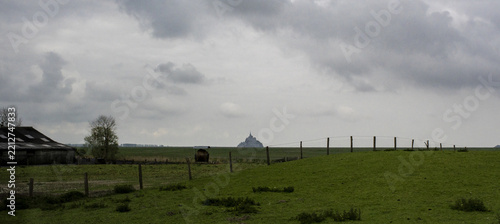 View of Mont Saint-Michel in the distance across green fields with a wooden fence and a rustic barn under a cloudy sky in Mont Saint-Michel, Normandy, France.