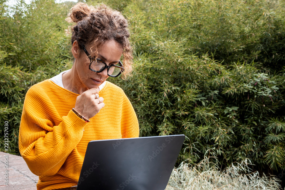 Fototapeta premium Woman in yellow sweater focuses on laptop while working remotely outdoors, thoughtful as she reads, researches and manages tasks in a peaceful garden with green foliage and plants.