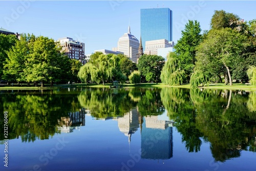 Boston skyline reflected in the tranquil pond of Boston Public Garden, surrounded by lush green trees and historic downtown buildings under a clear blue sky.