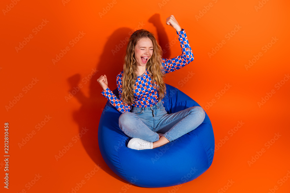 Fototapeta premium Cheerful young woman celebrating a moment of happiness while sitting on a vibrant blue bean bag against a bright orange background