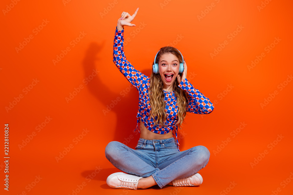 Fototapeta premium Excited young girl wearing stylish outfit sitting cross-legged and enjoying music with headphones on bright orange background