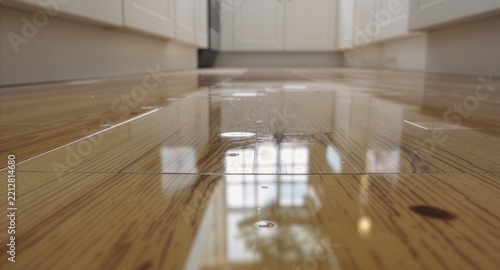 Low angle view of water leaking and pooling on a shiny wooden parquet floor in a white modern kitchen, representing a home plumbing accident, broken pipe emergency, or insurance damage.