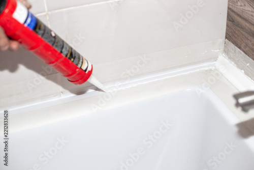 A worker uses a red caulking gun to apply a line of white silicone sealant along a clean bathroom tile corner. Residential bathtub restoration routine. Home improvement theme.