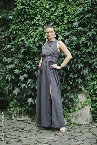 Woman stands in front of wall covered with green ivy. She wears long dark dress and looks straight forward. Her hair is styled and she wears accessories