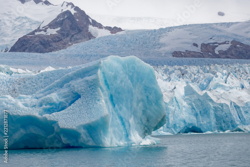 Massive cyan blue iceberg floating on cold lake waters near a melting glacier terminus against rugged snowy Patagonian mountain peaks.