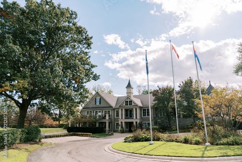 Historic mansion with flagpoles beside curved driveway, framed by tall trees, manicured lawn, flowering shrubs, and bright summer sky, showing elegant residential architecture and formal grounds