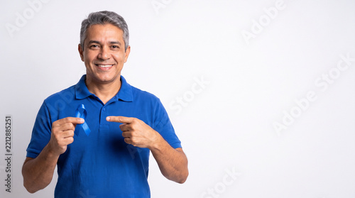 Middle-aged smiling man holding and pointing at a blue ribbon for prostate cancer awareness against a white background