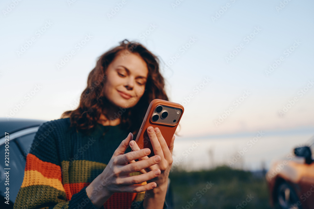 Obraz premium Young woman checks her phone outdoors at sunset, wearing a colorful sweater. She smiles softly as a candid moment unfolds in natural light.
