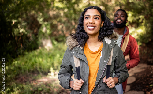 A joyful Black couple enjoys a hike in a lush green forest. The woman, wearing a warm jacket, leads the way with a bright smile, while the man follows closely behind, both embracing the beauty of natu