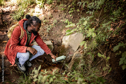 A man crouches by a natural spring in a forest, filling a water bottle. He wears a red jacket and has a backpack. The scene is serene, surrounded by greenery and sunlight filtering through the trees.