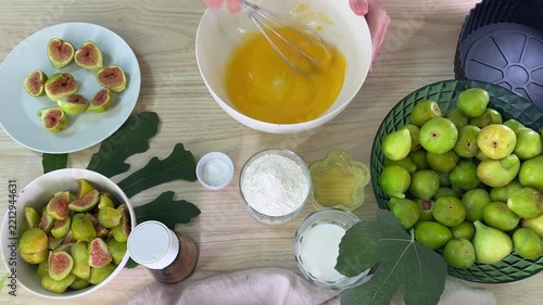a person mixes yellow batter in a white bowl, surrounded by green figs in a basket, sliced figs on a plate, and bowls of white powder and liquid