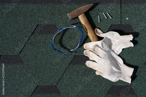 Roofing tools including a hammer, safety glasses, work gloves, and nails lying on green hexagonal asphalt shingles. Top-view flat lay.