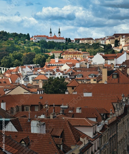 Elevated view across the historic red rooftops of Malá Strana toward Strahov Monastery and the Basilica of the Assumption in Prague, Czech Republic, beneath dramatic clouds.