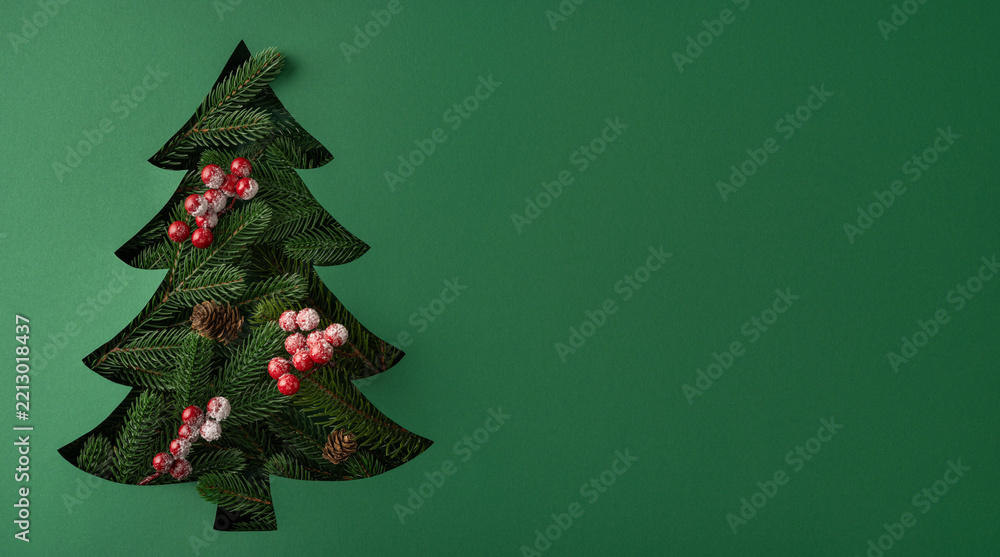 Fototapeta premium Minimal green Christmas tree cutout with frosted berries and pinecones for New Year greeting on banner green background