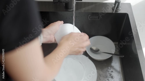 Woman scrubs and rinses bowl with sponge under running water while other dishes remain in sink. Everyday tableware cleaning and kitchen hygiene