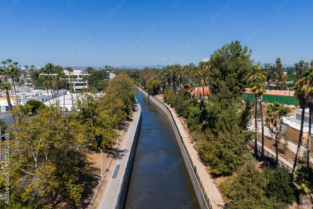 Fototapeta premium Ballona Creek concrete flood control channel flows beside a paved bicycle path and trees under a clear sky in Culver City, Los Angeles County, California.