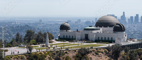 Griffith Observatory sits on the slope of Mount Hollywood in Griffith Park, with visitors on the grounds and the downtown Los Angeles city skyline visible under hazy daylight.